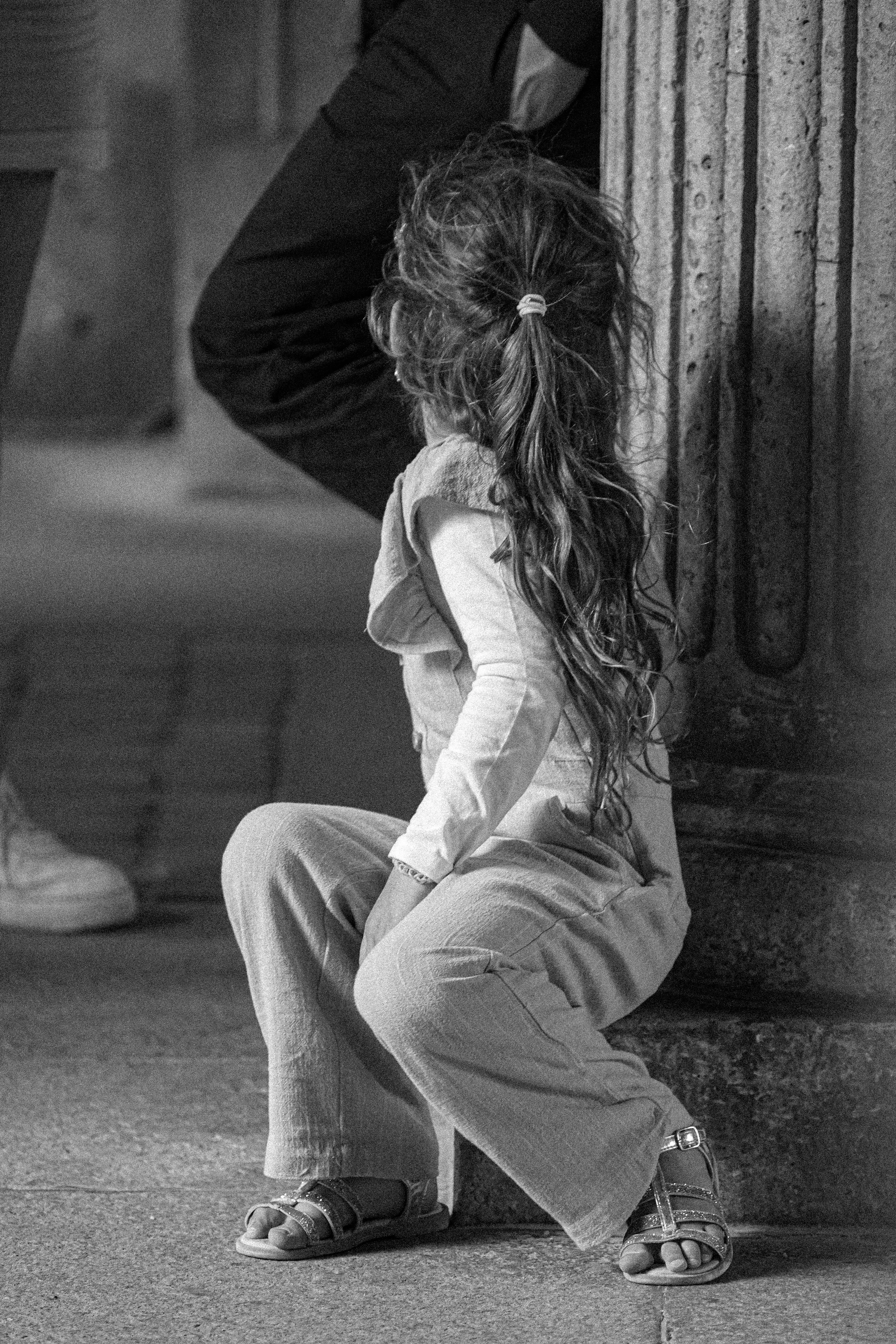 Black and white photograph of a young girl in wide trousers standing beside a stone column, seen from behind