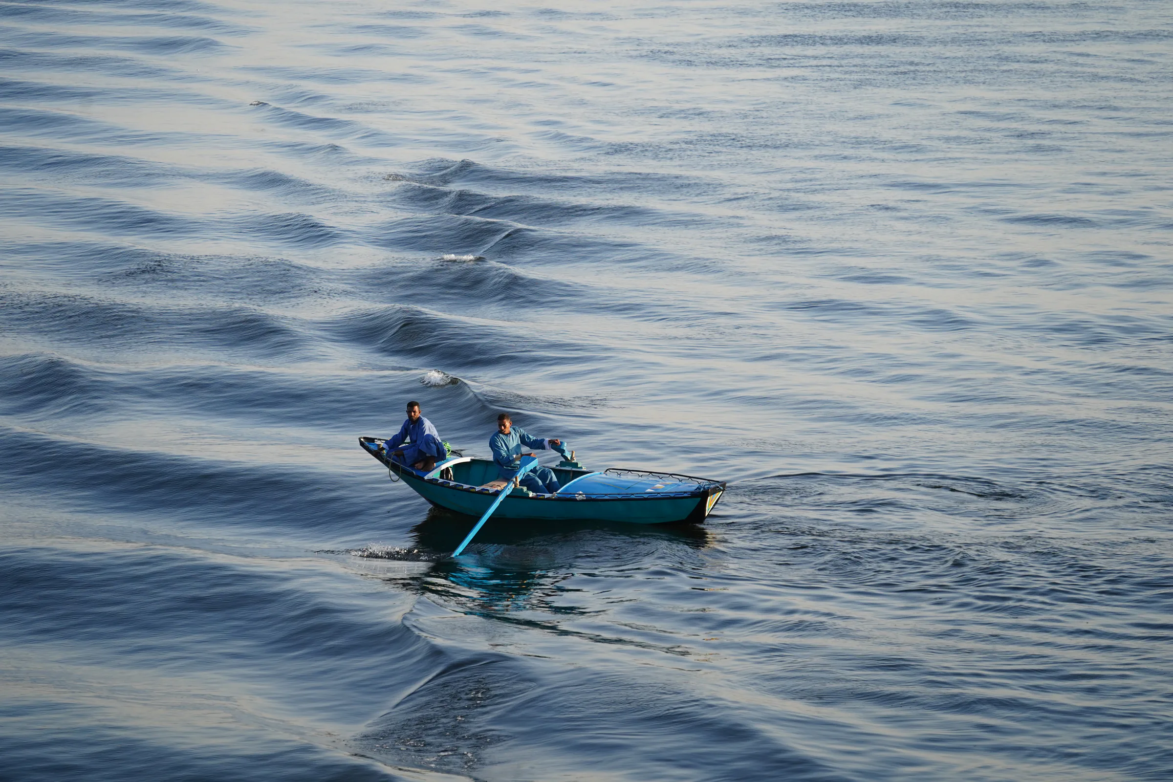 Two men in blue robes crossing calm water in a small blue boat
