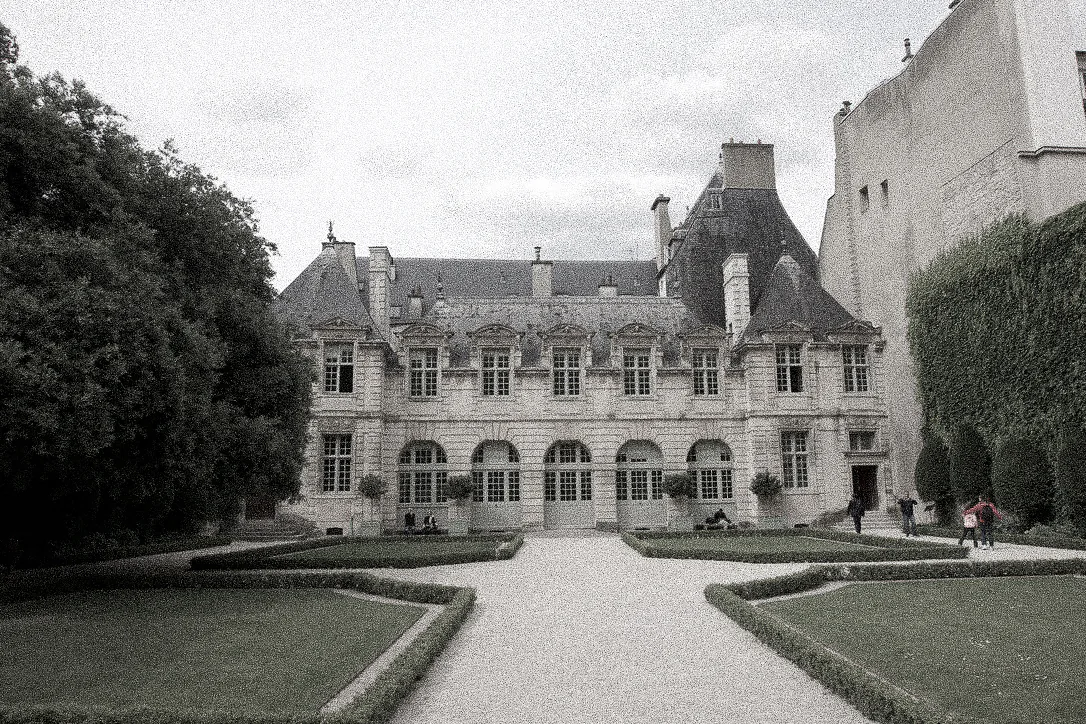 A mansard-roofed mansion behind a formal garden of clipped hedges, in black and white