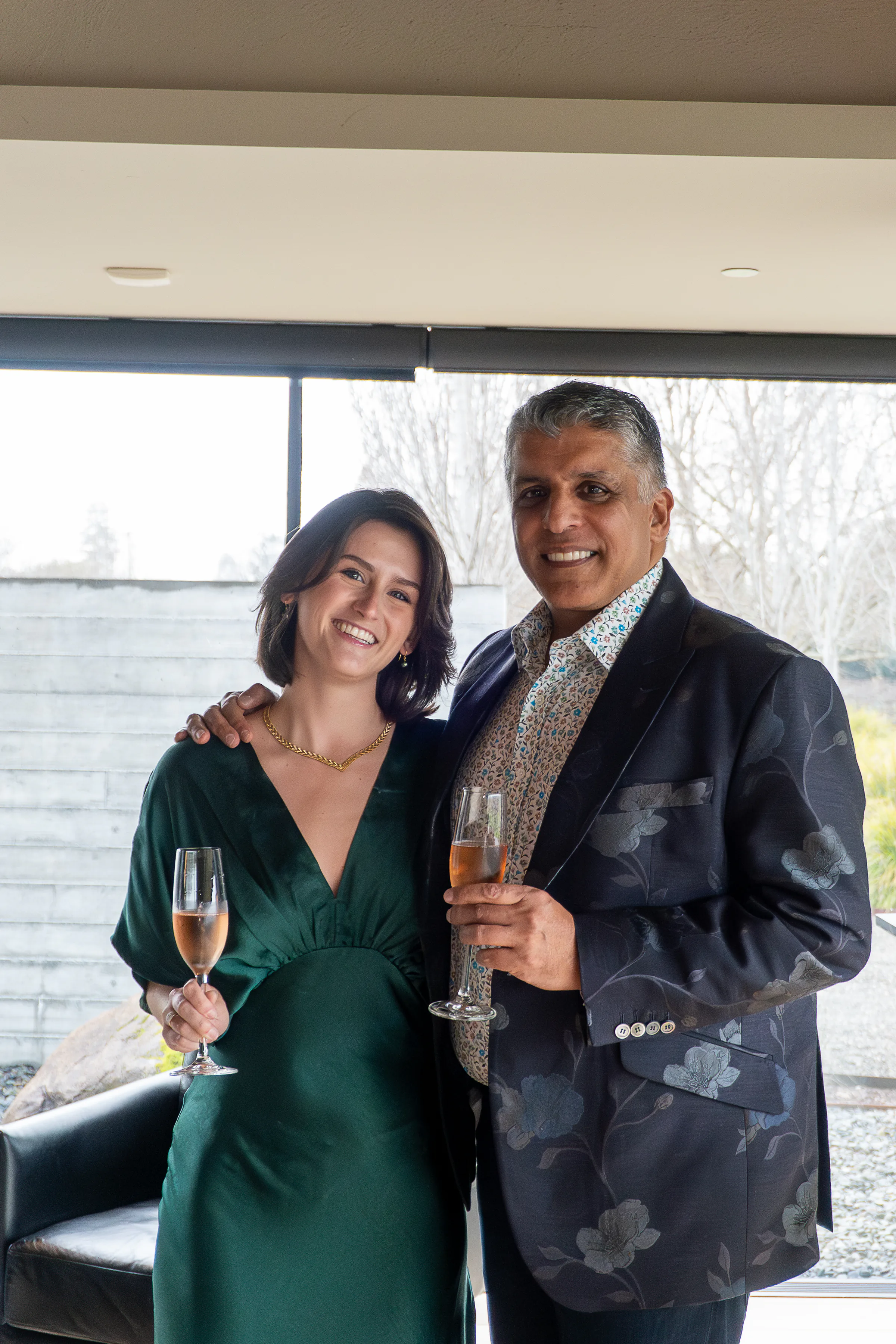 A woman in a green dress and a man in a floral blazer holding champagne glasses beside a window